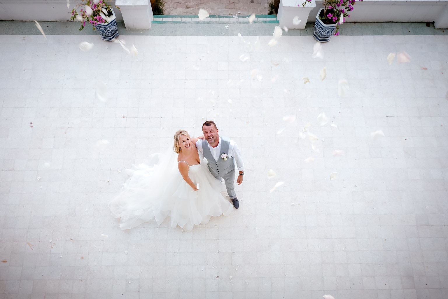 A bride and groom looking up at the camera with smiles, surrounded by falling petals, on a tiled outdoor surface.