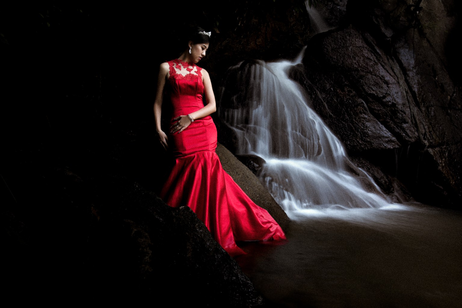 A woman in a red gown stands near a waterfall, surrounded by dark rocks and water.