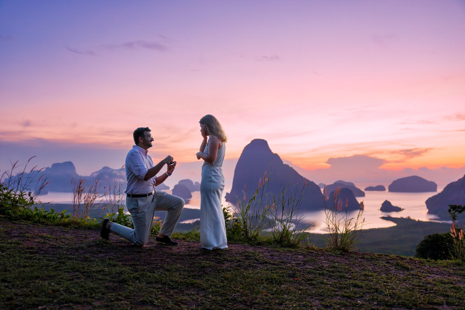 A romantic proposal scene at sunset, featuring a man kneeling with a ring in hand, and a woman in a white dress, looking surprised, against a backdrop of mountains and water.