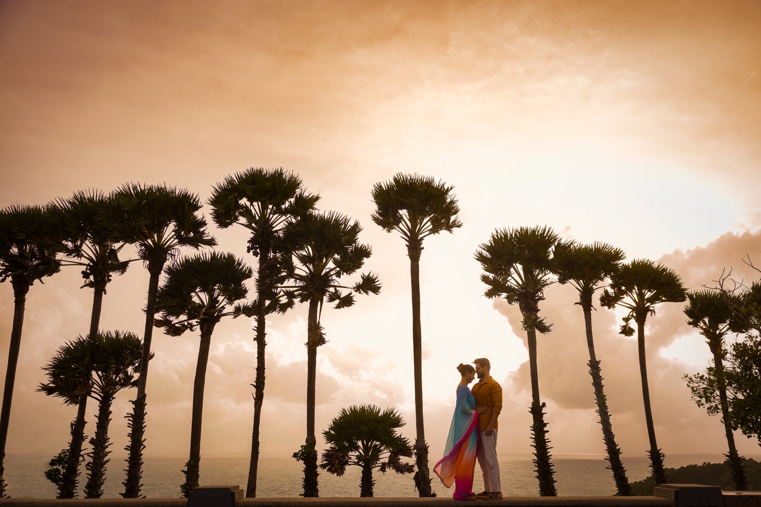 Silhouetted couple standing together at Promthep Cape, Phuket, with palm trees and a dramatic sunset in the background.