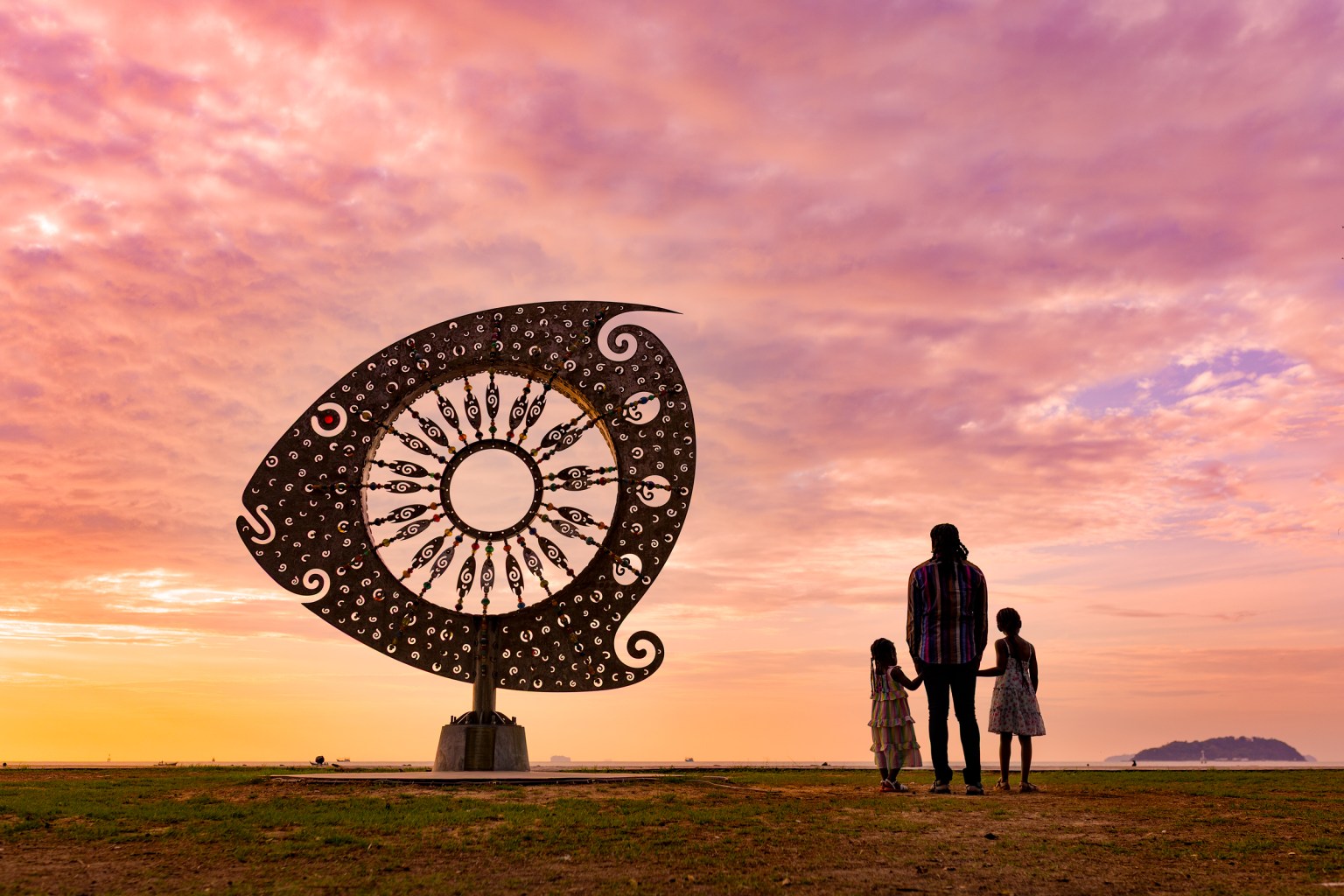 Silhouette of a family standing near a large decorative fish sculpture against a colorful sunrise sky.