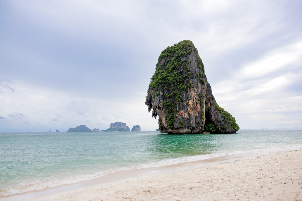 Scenic view of a limestone rock formation in the sea at Railay Beach, Krabi, Thailand, with a sandy shore and cloudy sky.