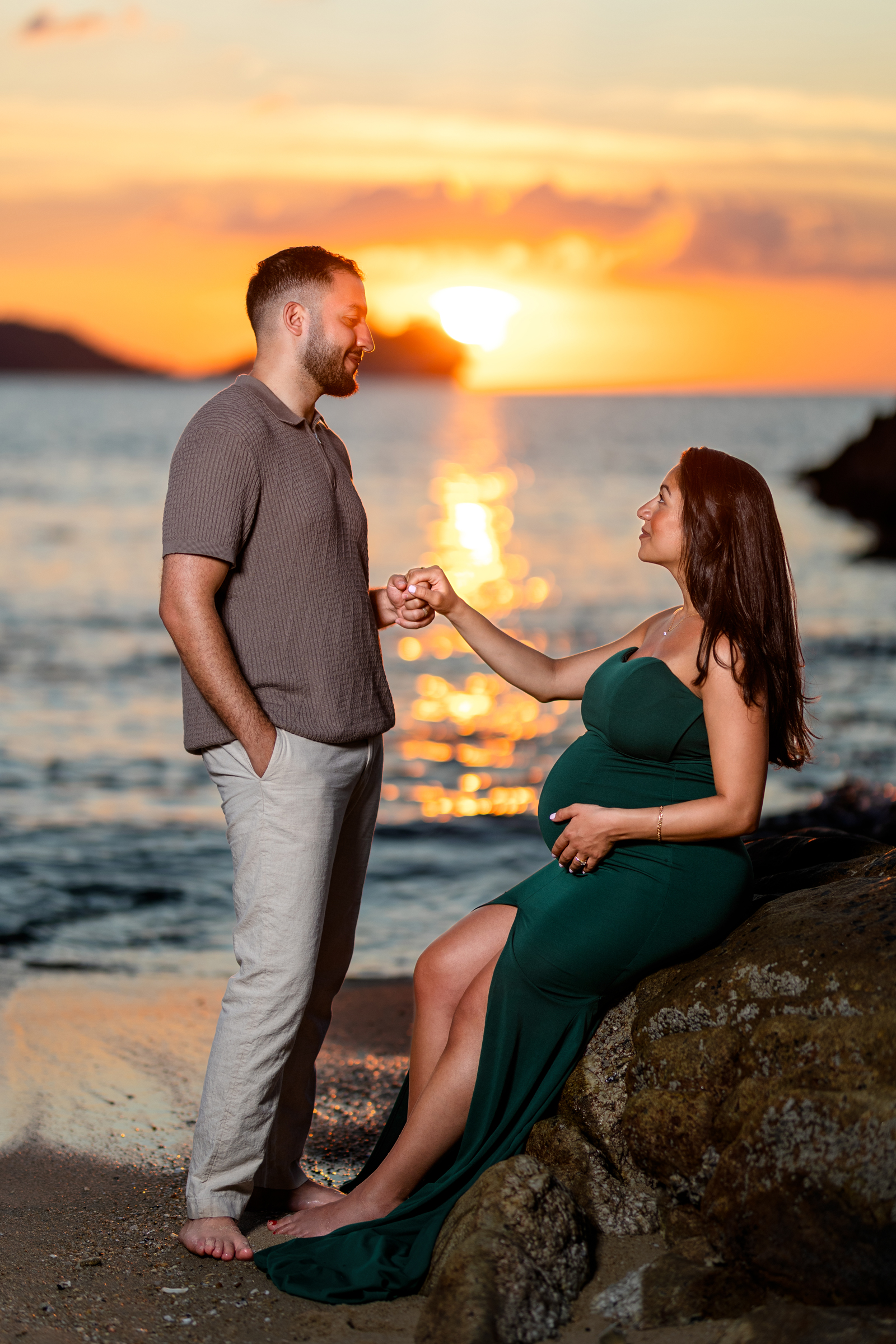 A couple embracing at the beach during sunset, with the pregnant woman in a green dress and the man in a casual shirt, both looking at each other.