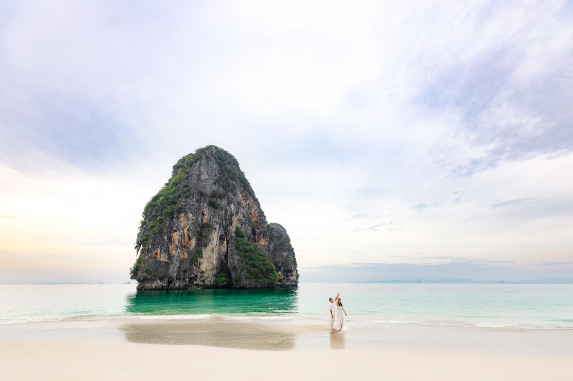 A couple dancing on the beach at Railay, Krabi, Thailand, with a large rock formation in the background and a calm sea.
