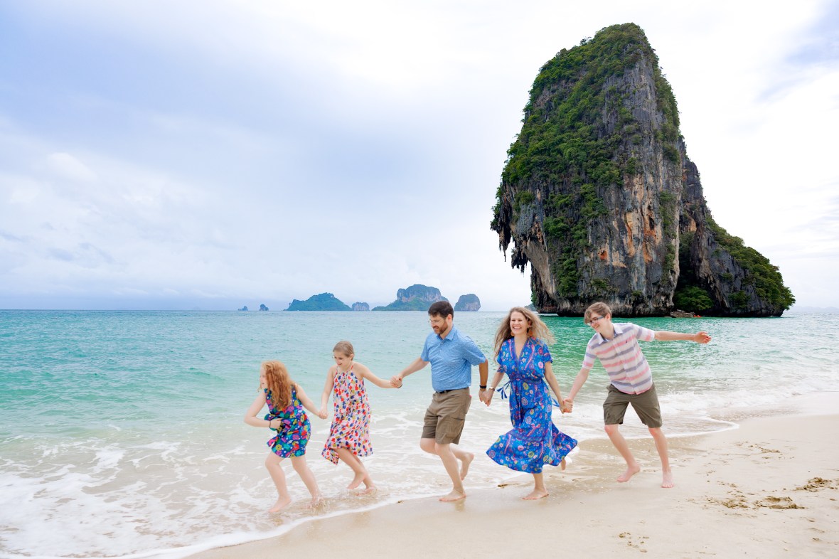 A family enjoying a playful moment at the beach, holding hands and splashing in the water, with a large rock formation in the background.