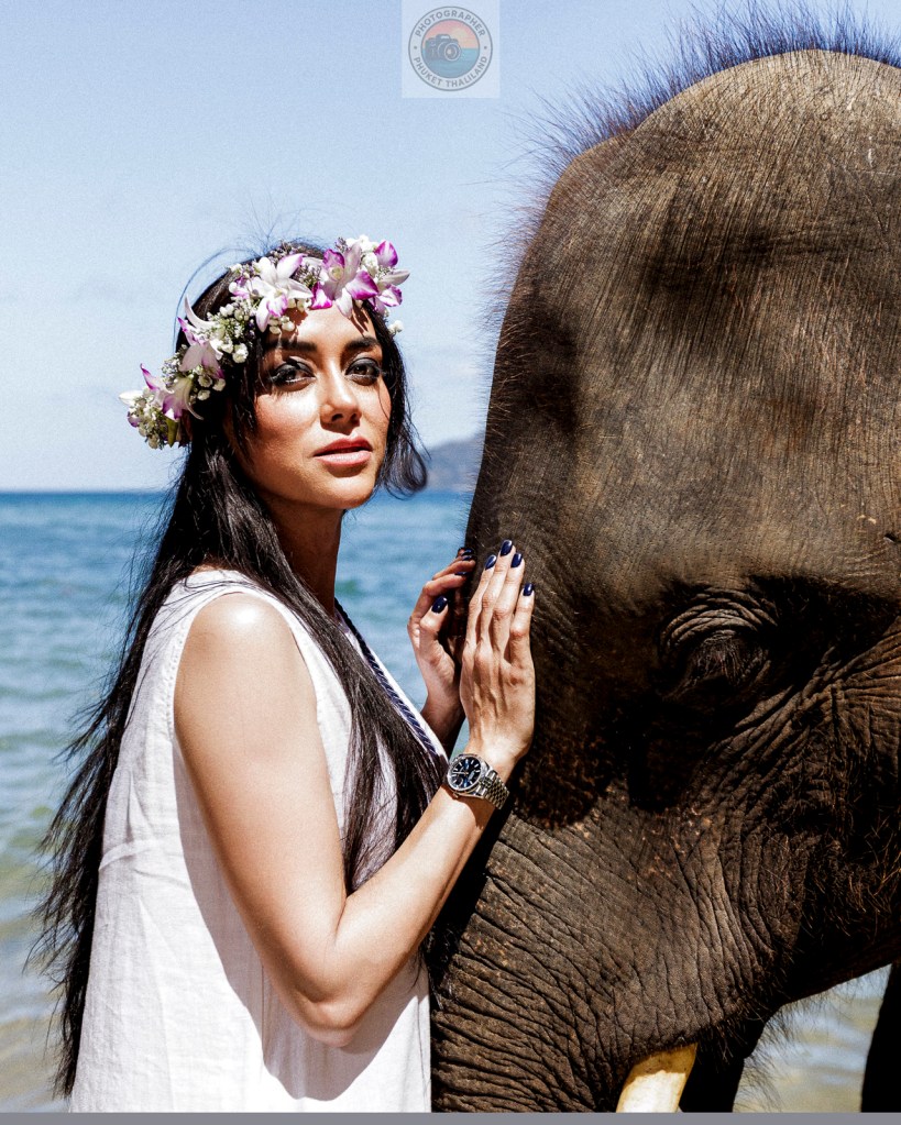 A woman with a floral crown poses next to an elephant at the beach, with the ocean in the background.