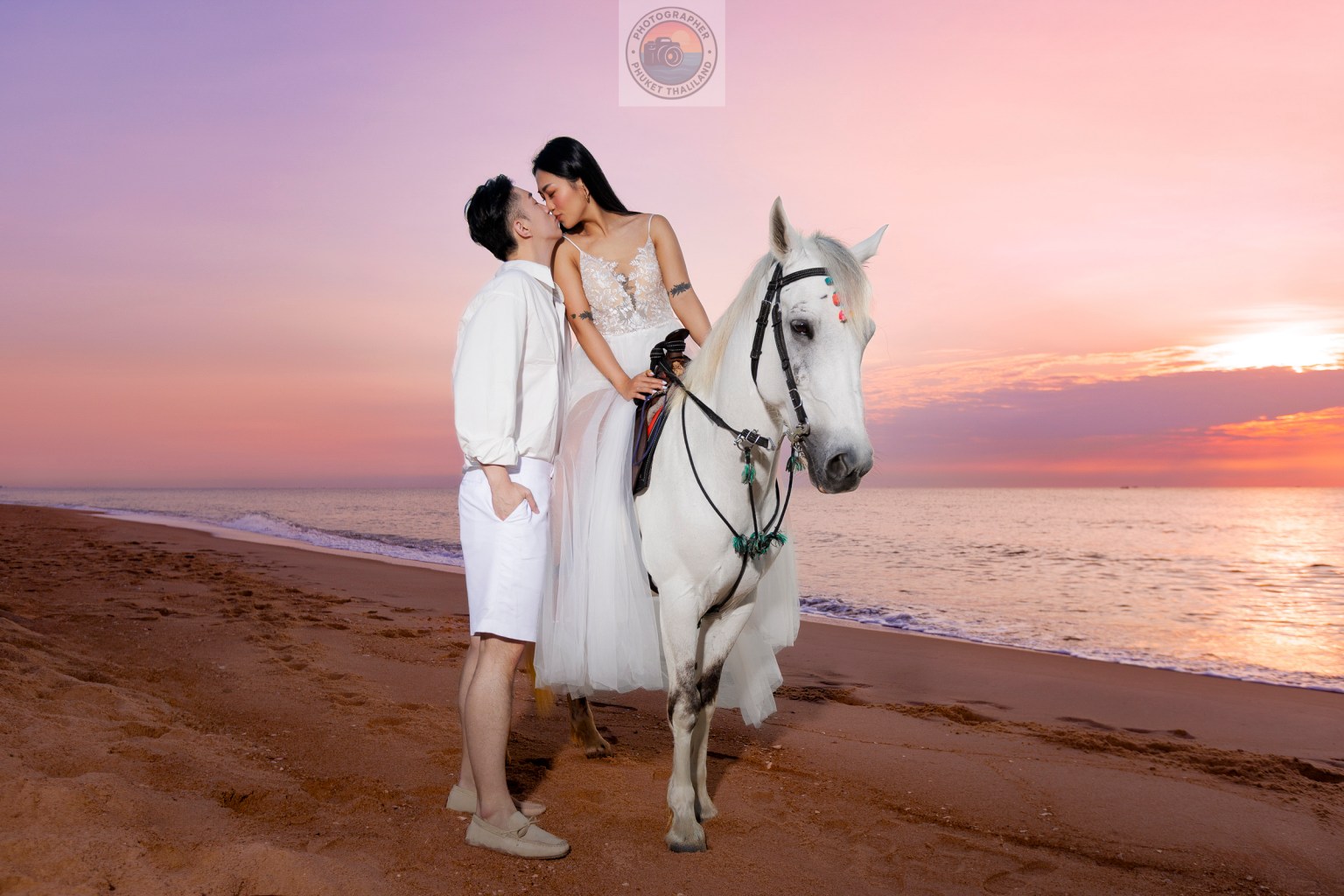 A man and a woman share a romantic moment on a beach at sunset, with the woman riding a white horse.