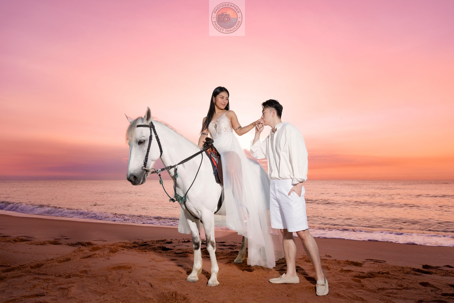A couple on a beach at sunset, with the woman wearing a white dress and sitting atop a white horse, while the man leans in to kiss her hand.