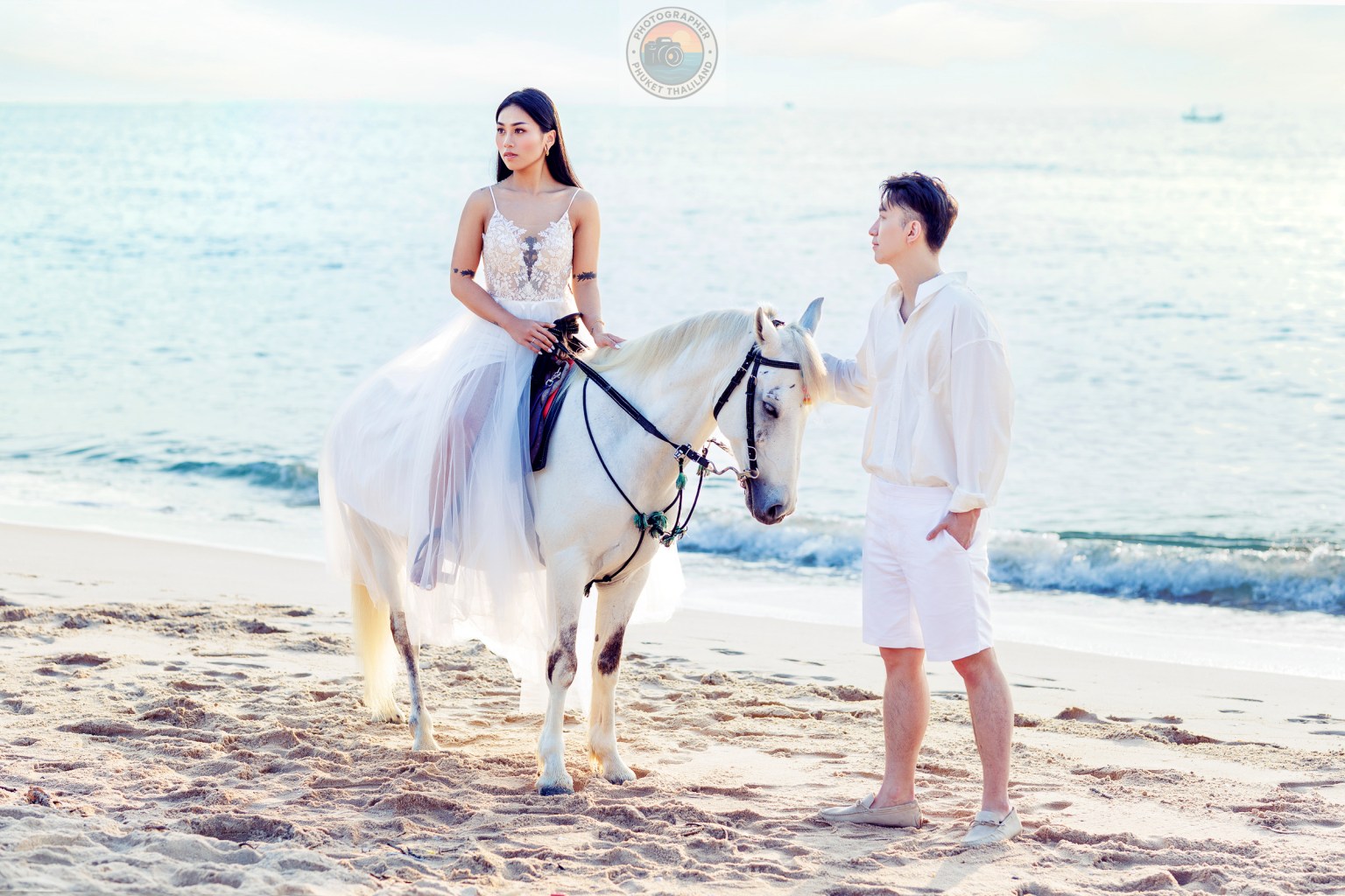 A woman in a white dress sitting on a white horse on a beach, with a man standing beside her, both looking towards the ocean.