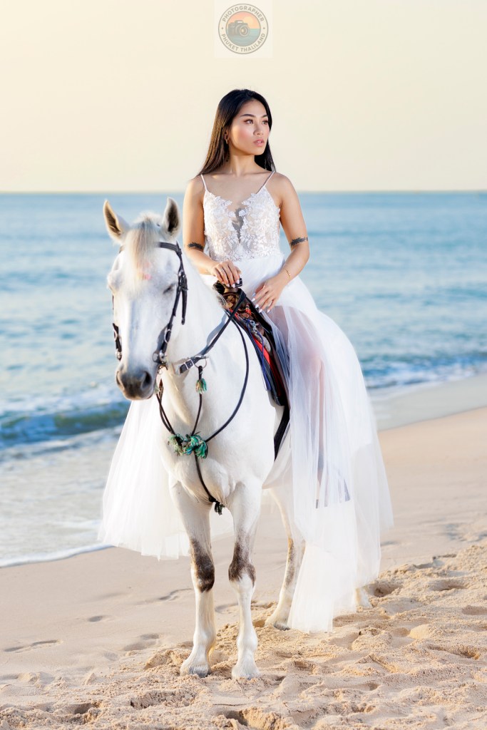 A woman in a beautiful white gown riding a white horse on a sandy beach with a calm ocean in the background.