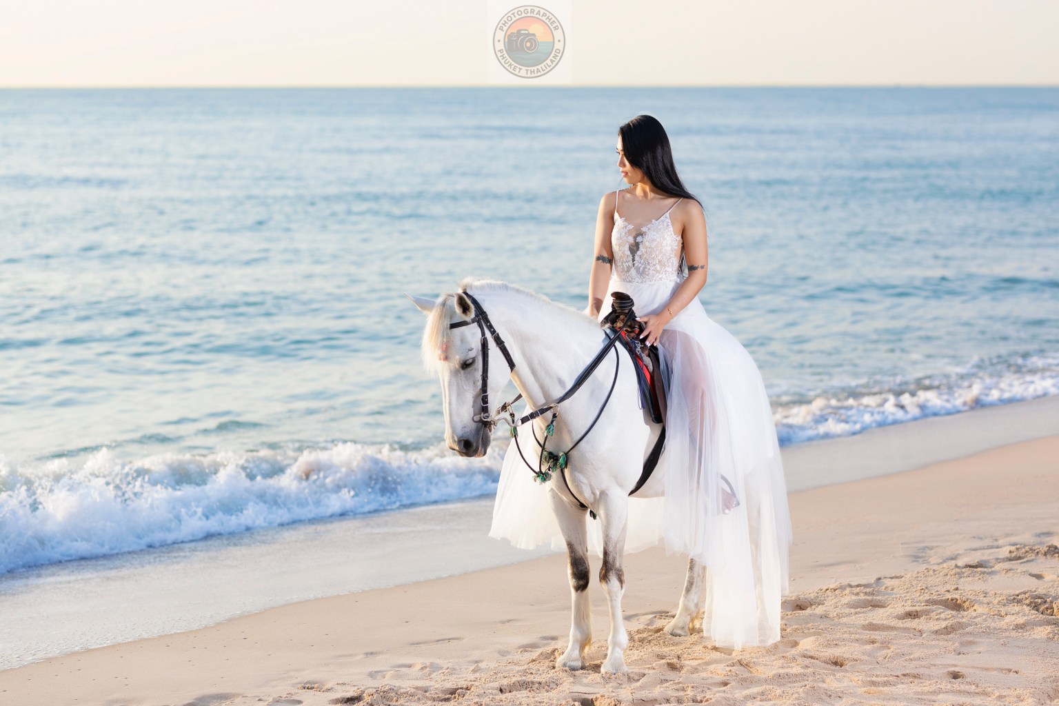 A woman in a white dress riding a white horse on a beach, with the ocean in the background and gentle waves lapping at the shore.
