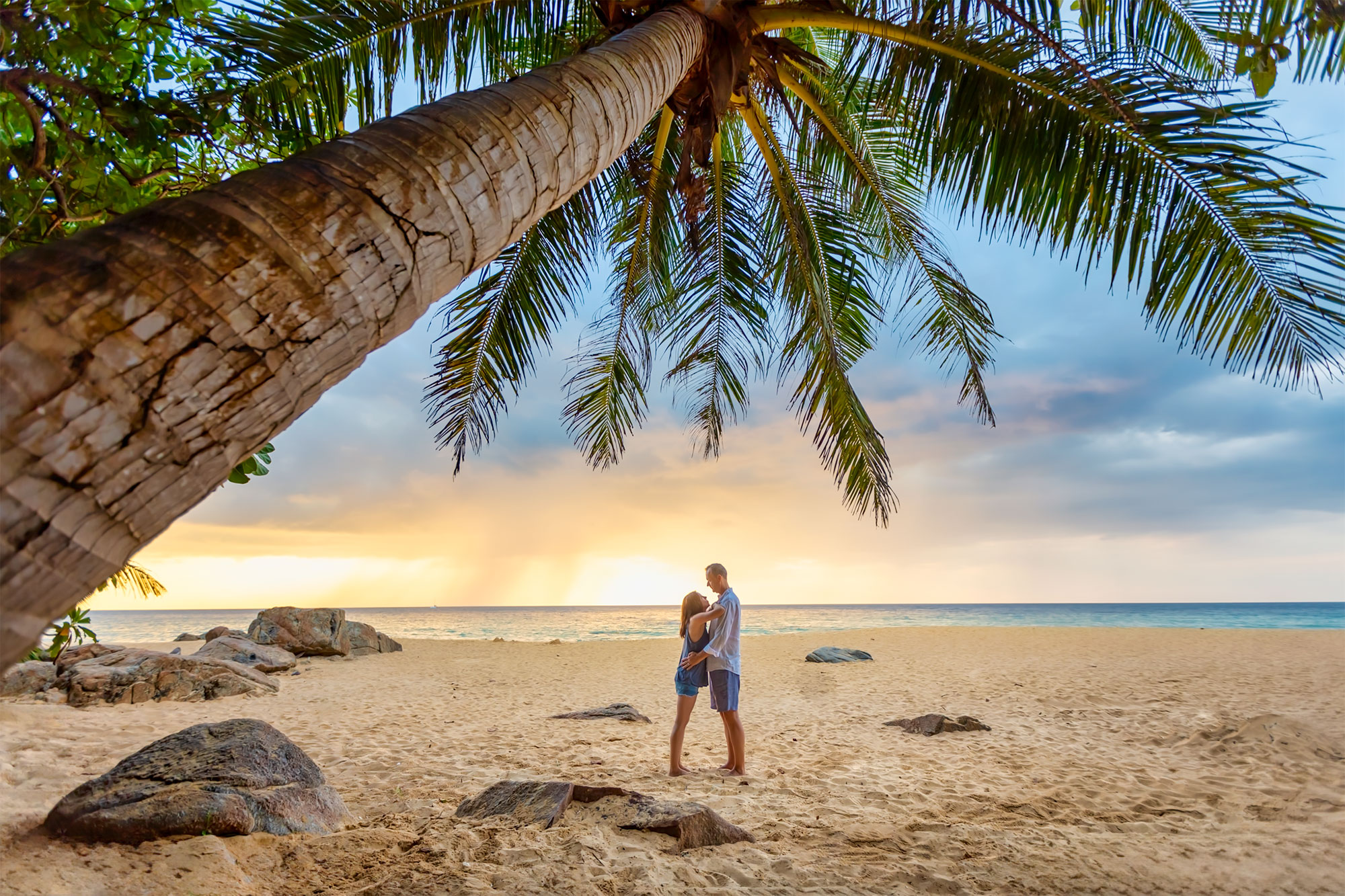 A couple embracing on a sandy beach at sunset, framed by palm trees and rocky terrain.