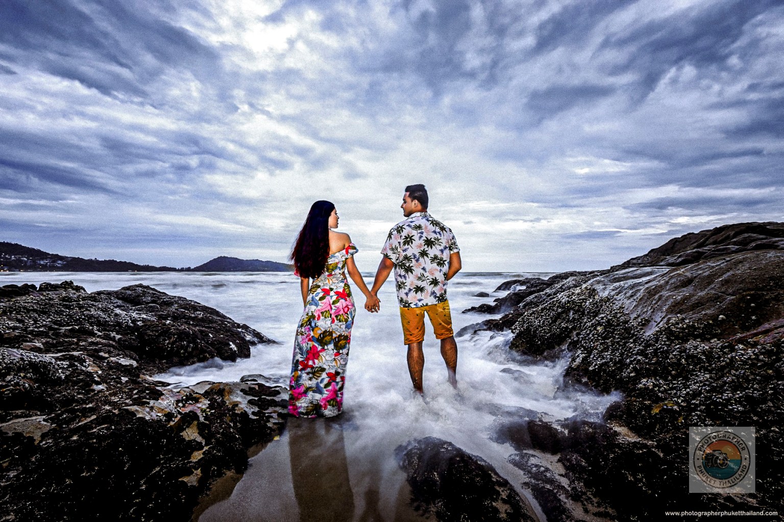 A couple holding hands while standing on rocky beach terrain, facing the ocean under a cloudy sky.