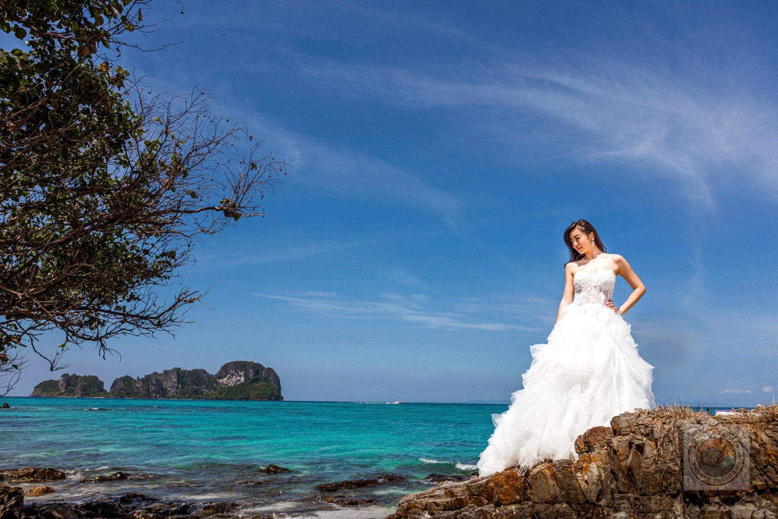 A bride in a white wedding dress stands on rocks by the sea, with a clear blue sky and distant islands in the background.