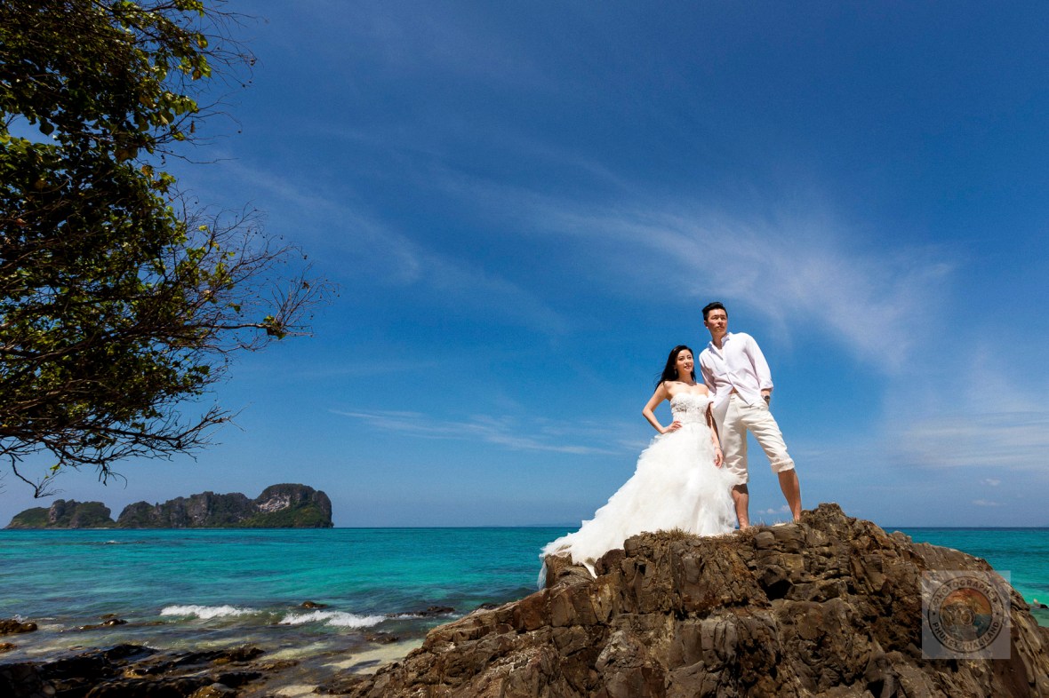 A couple in wedding attire poses on a rocky outcrop by the beach, with turquoise waters and a clear blue sky in the background at bamboo island phi phi krabi.
