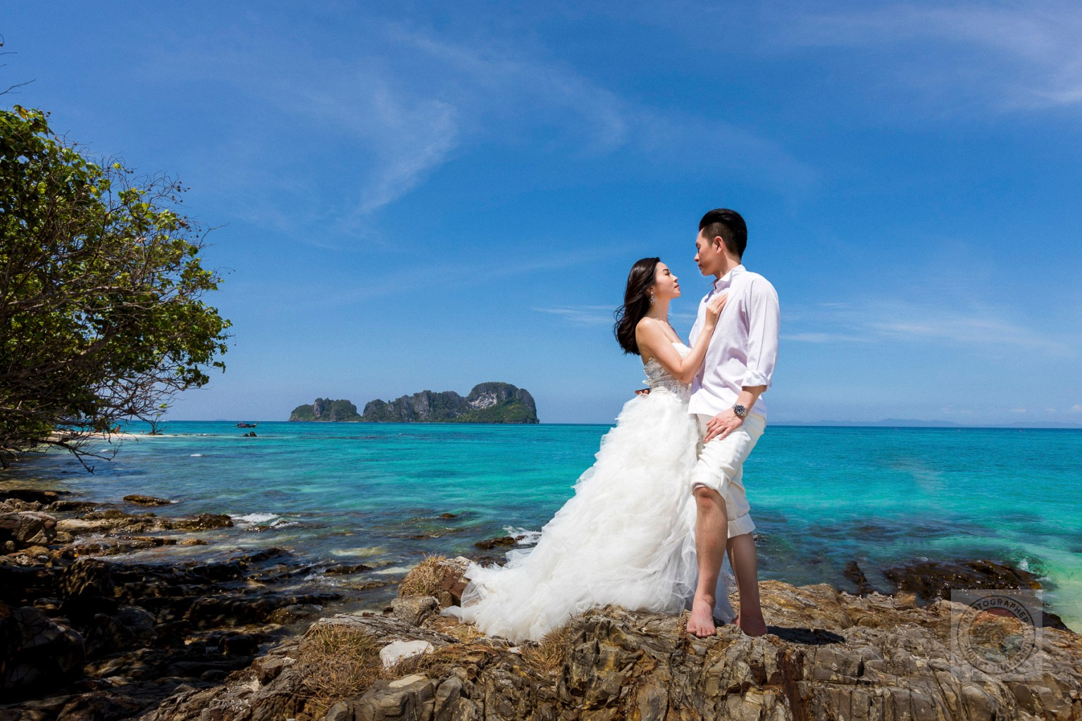 A couple poses on rocky terrain near the shore, with a bright blue ocean and islands in the background. The woman is wearing a flowing white wedding dress, while the man is dressed in a casual white shirt and light pants.