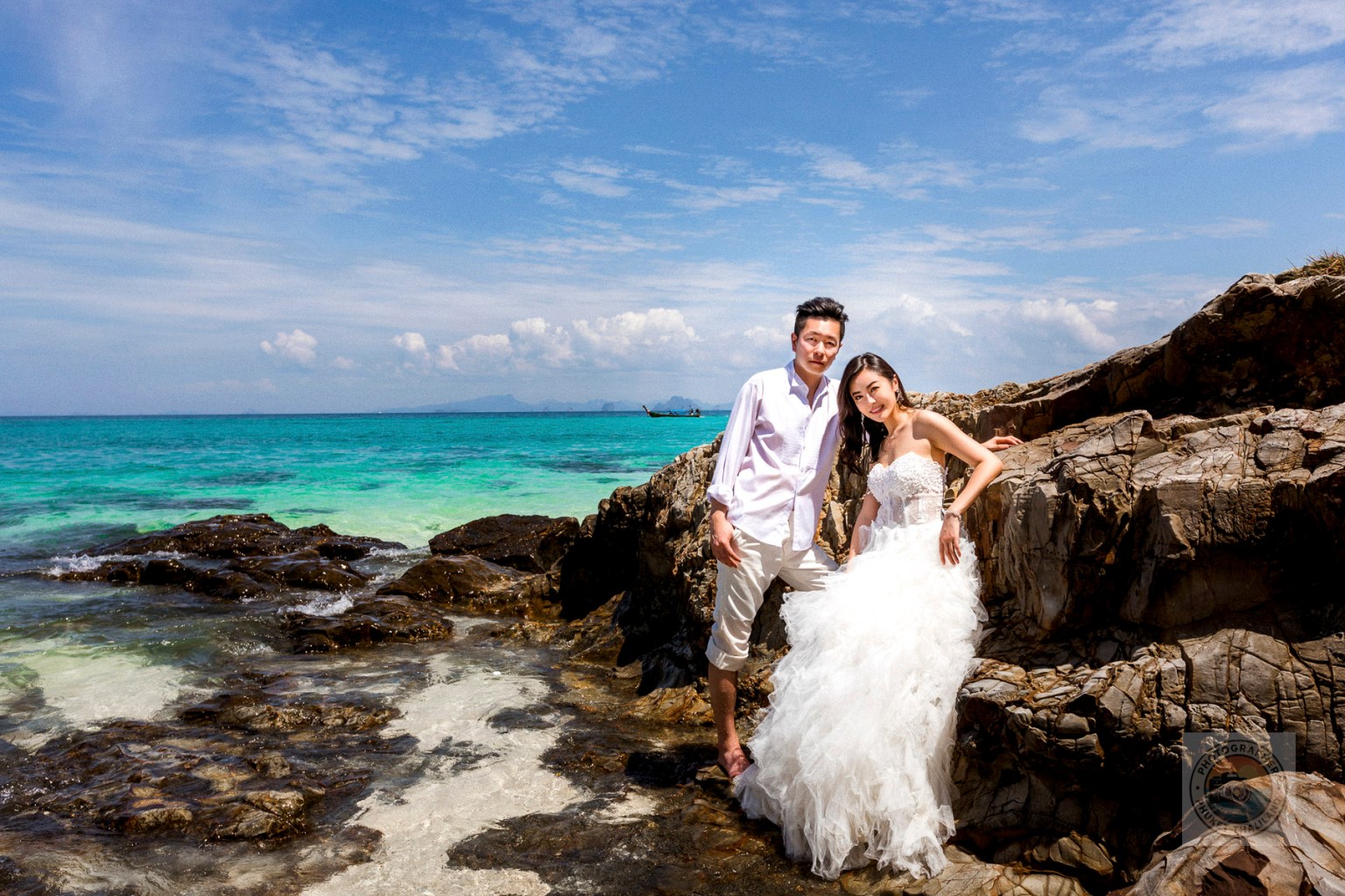 A couple posing happily on the rocks by the beach, with crystal clear turquoise waters and a bright blue sky in the background.