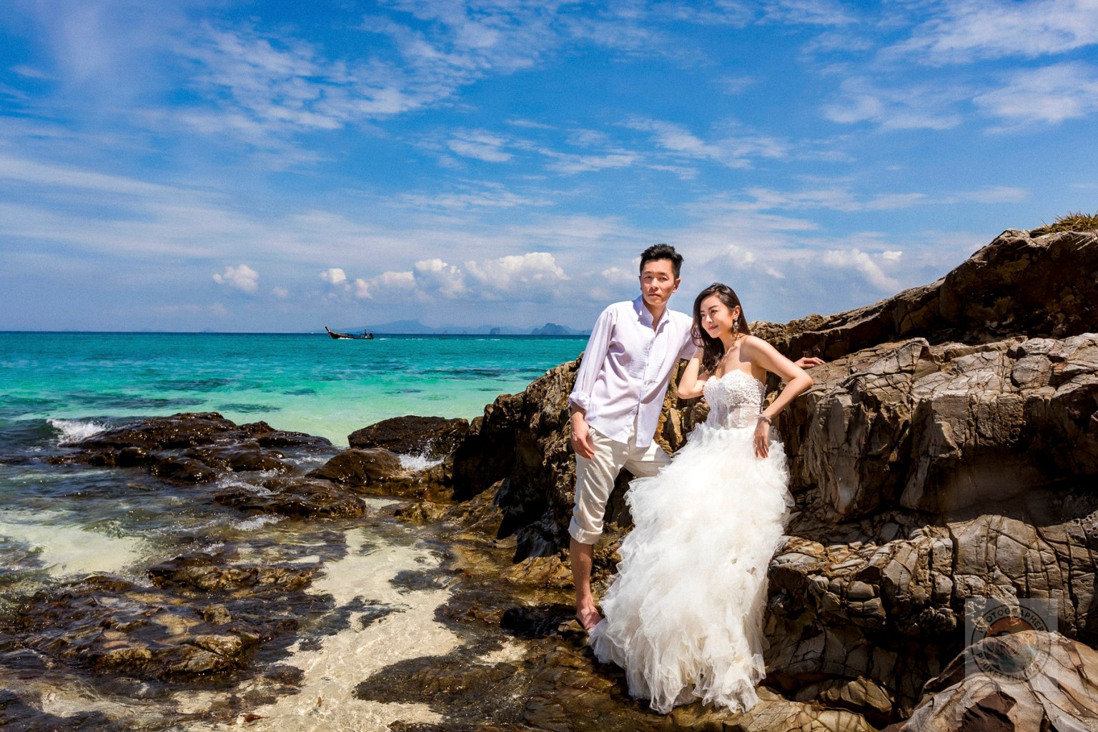 A couple poses on rocky beach terrain in Krabi, Thailand, with vibrant turquoise waters and a blue sky in the background during a pre-wedding photoshoot.