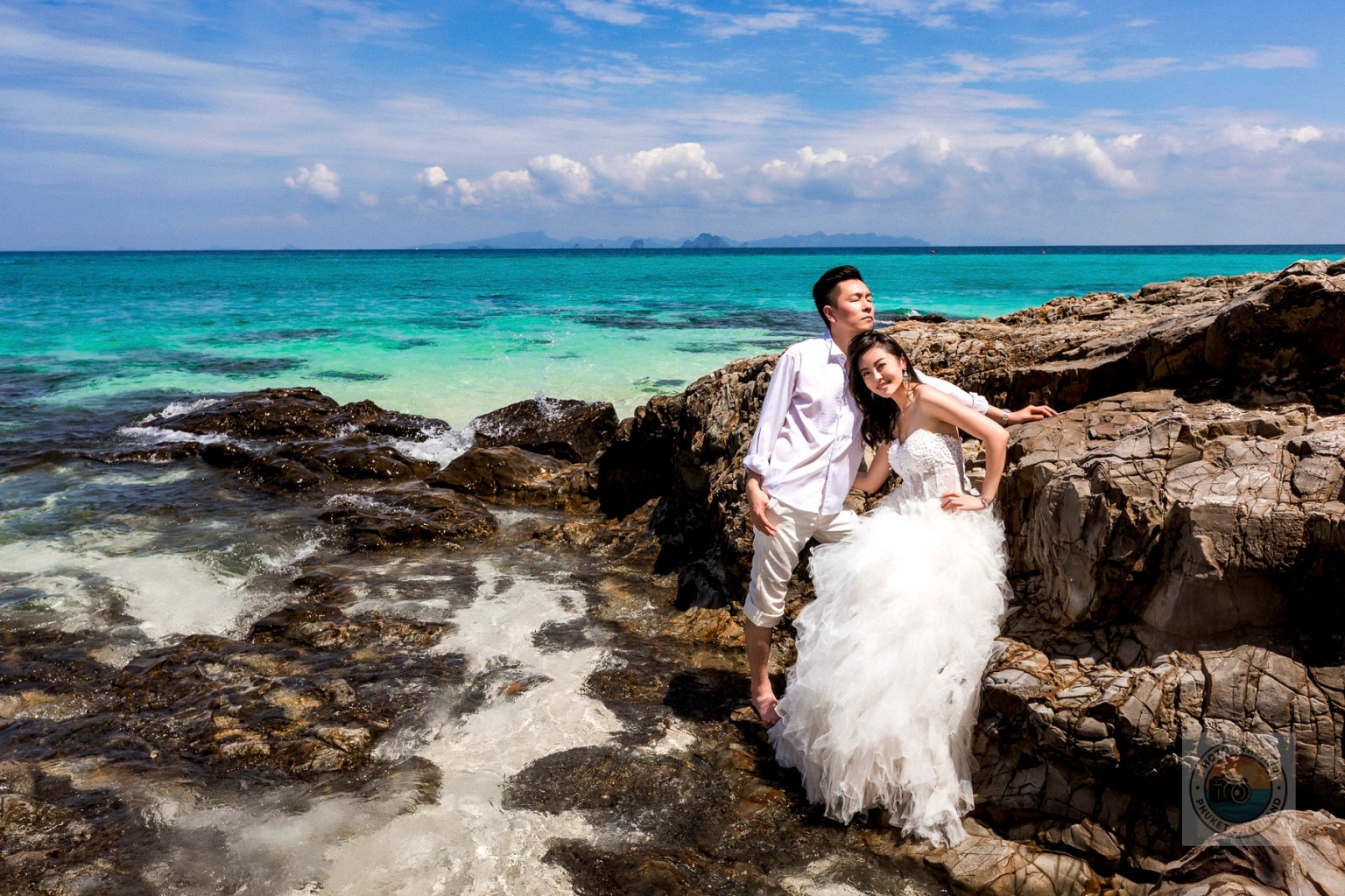 A couple in wedding attire poses on a rocky beach with turquoise water and blue sky in the background.