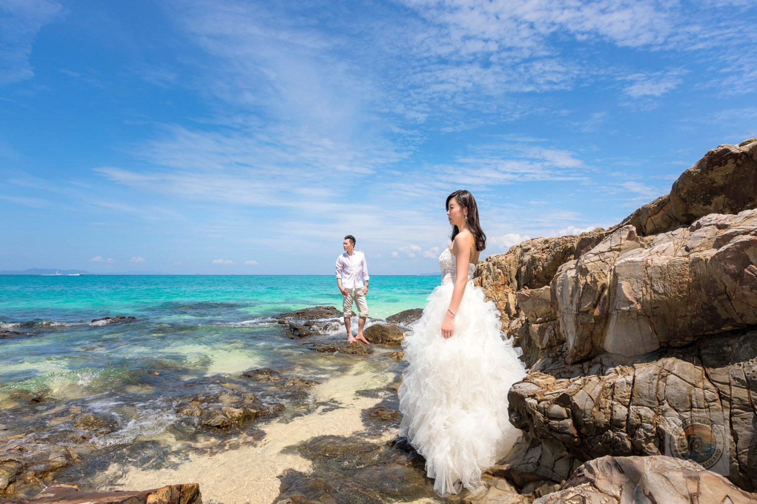 Couple posing for a pre-wedding photoshoot on a rocky beach with turquoise water and a blue sky.