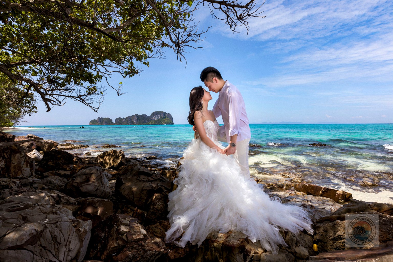 Couple in formal attire embracing on rocky beach with turquoise water and distant islands in the background.