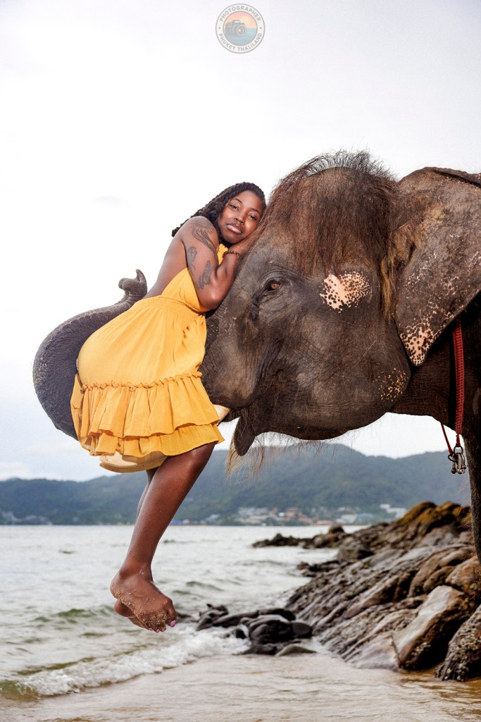 A woman in a yellow dress playfully poses while hugging an elephant on the shore, with a scenic background of water and hills.
