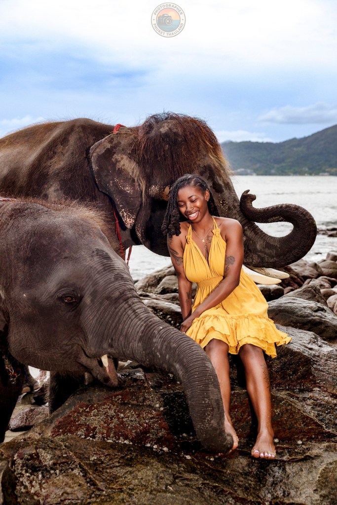 A woman in a yellow dress interacts happily with two elephants by the shore, surrounded by rocks and the sea under a cloudy sky.