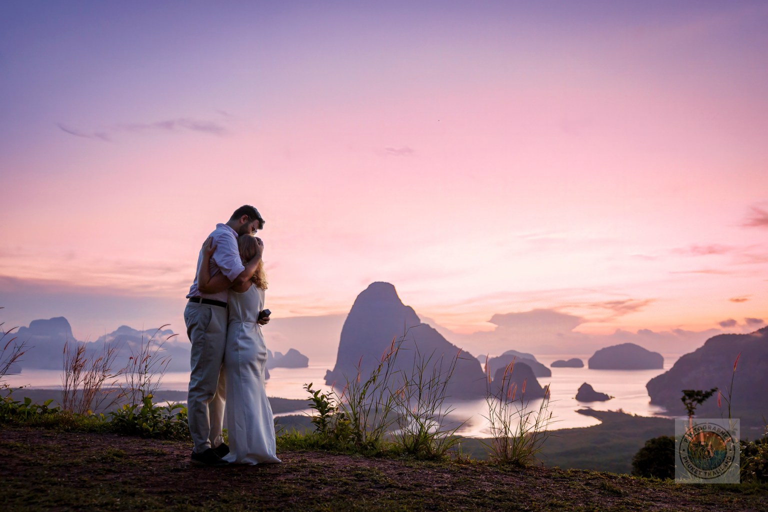 A couple embraces in a romantic pose against a picturesque sunset backdrop with mountains and water in the distance.