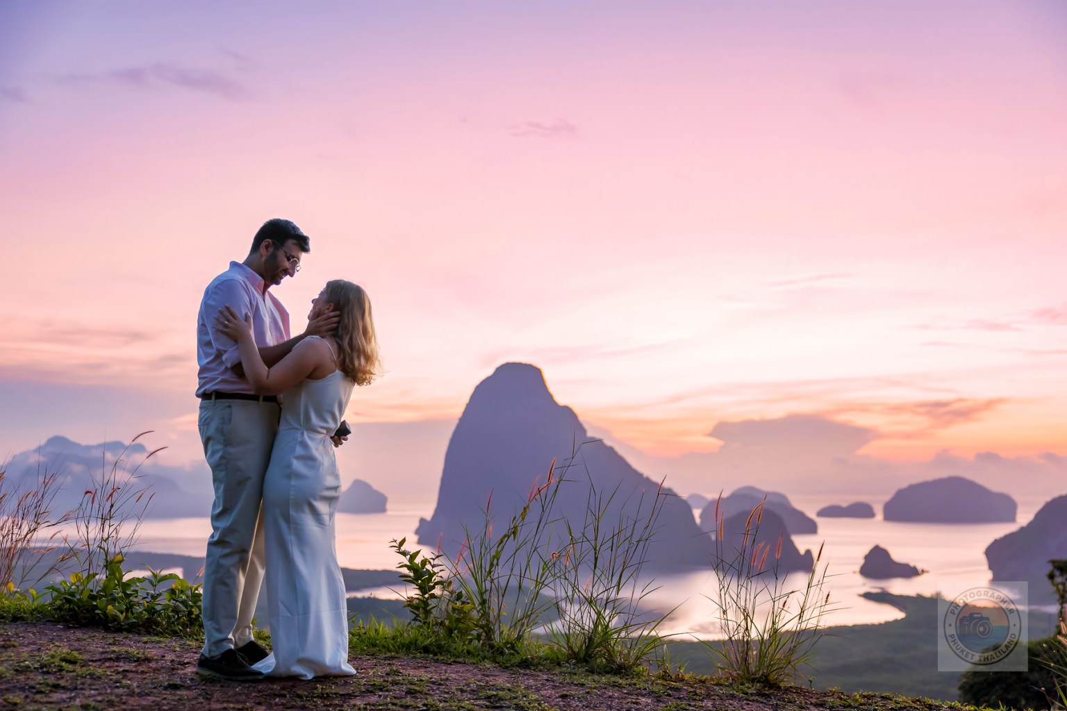 A couple embracing on a scenic overlook at sunset, with mountains and water in the background.