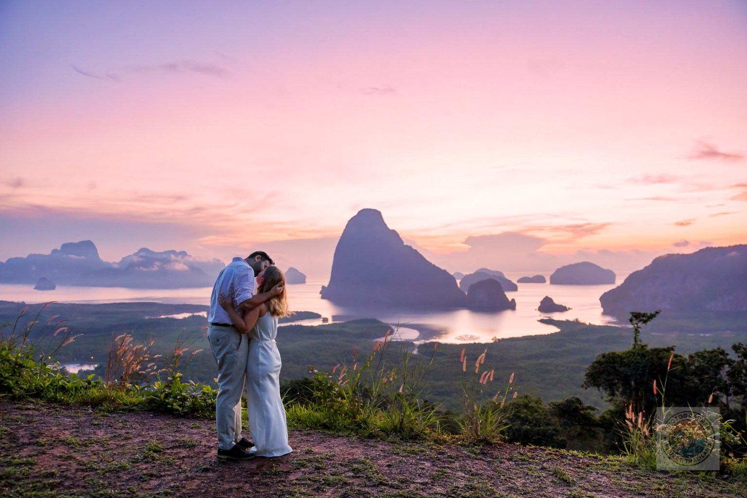 A couple embracing on a hillside with a scenic view of a bay at sunset, featuring dramatic rock formations and a colorful sky.