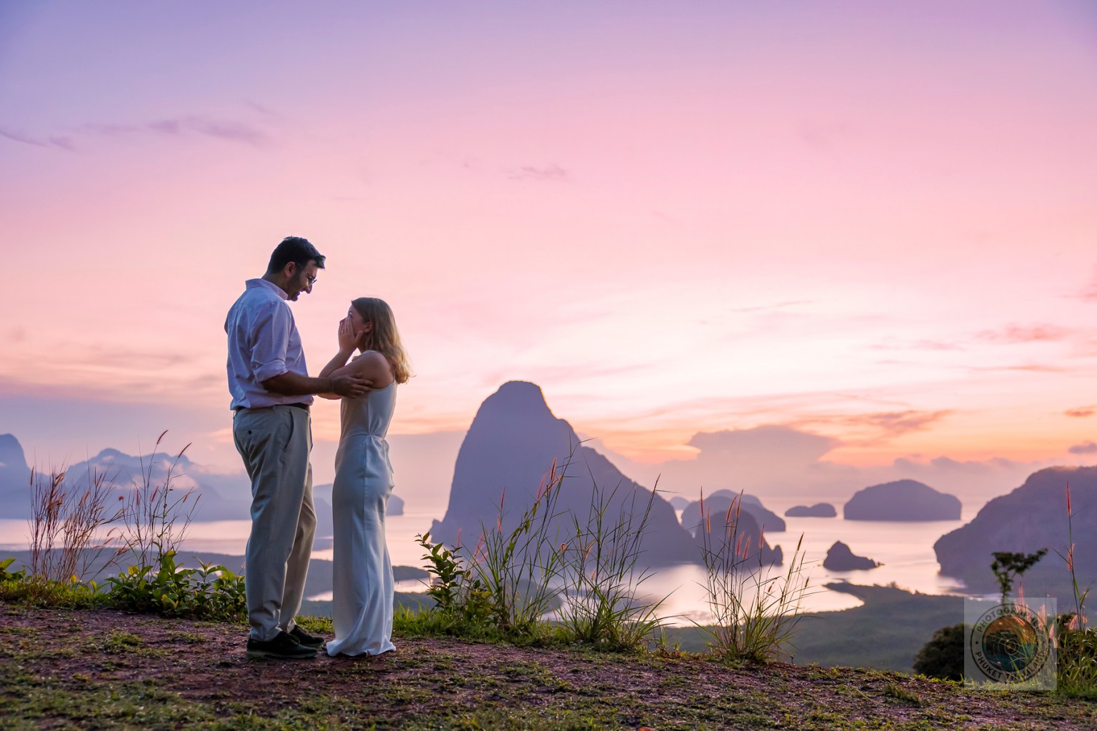 A couple embracing each other on a hilltop during sunset, with a scenic view of islands and the ocean in the background.