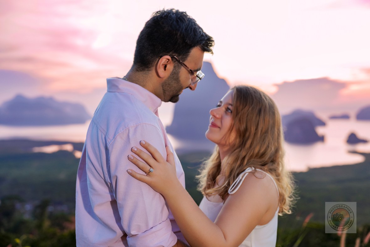 A romantic scene featuring a man and a woman gazing into each other's eyes against a backdrop of a colorful sunrise over a scenic landscape.