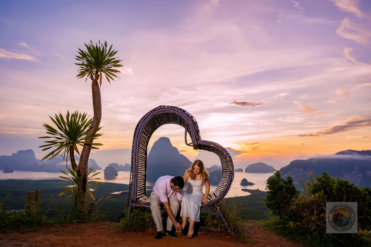 A couple sitting together in a heart-shaped bench with a scenic view of mountains and water during sunset.