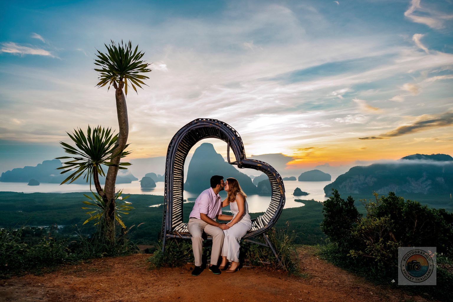 A couple kissing while sitting in a heart-shaped swing with a scenic view of mountains and water at sunset.
