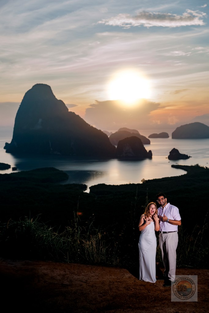 A couple joyfully celebrating their engagement or wedding at sunset, with stunning mountain and ocean views in the background.