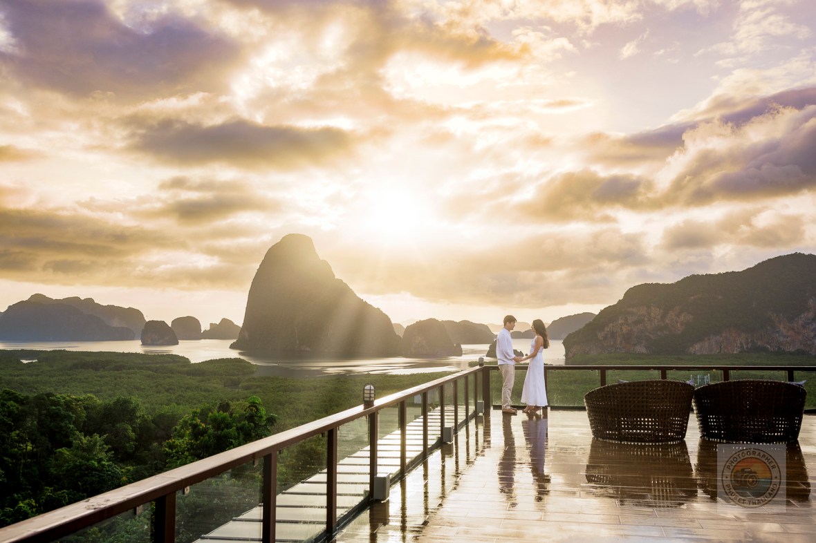 A couple stands together on a balcony at Samet Nangshe, overlooking the stunning limestone formations of Phang Nga Bay during sunset.