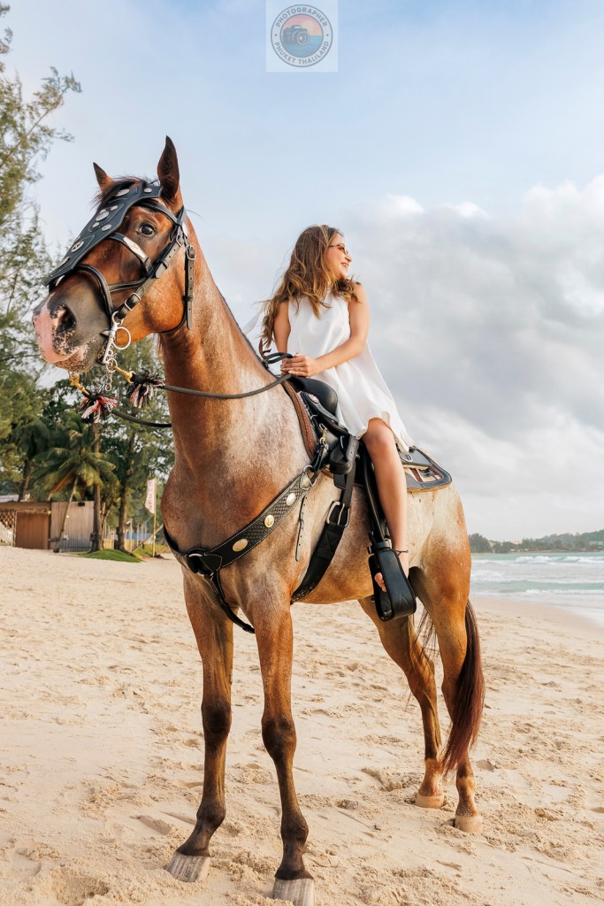 A woman in a white dress sits on a brown horse on a sandy beach with a cloudy sky in the background.