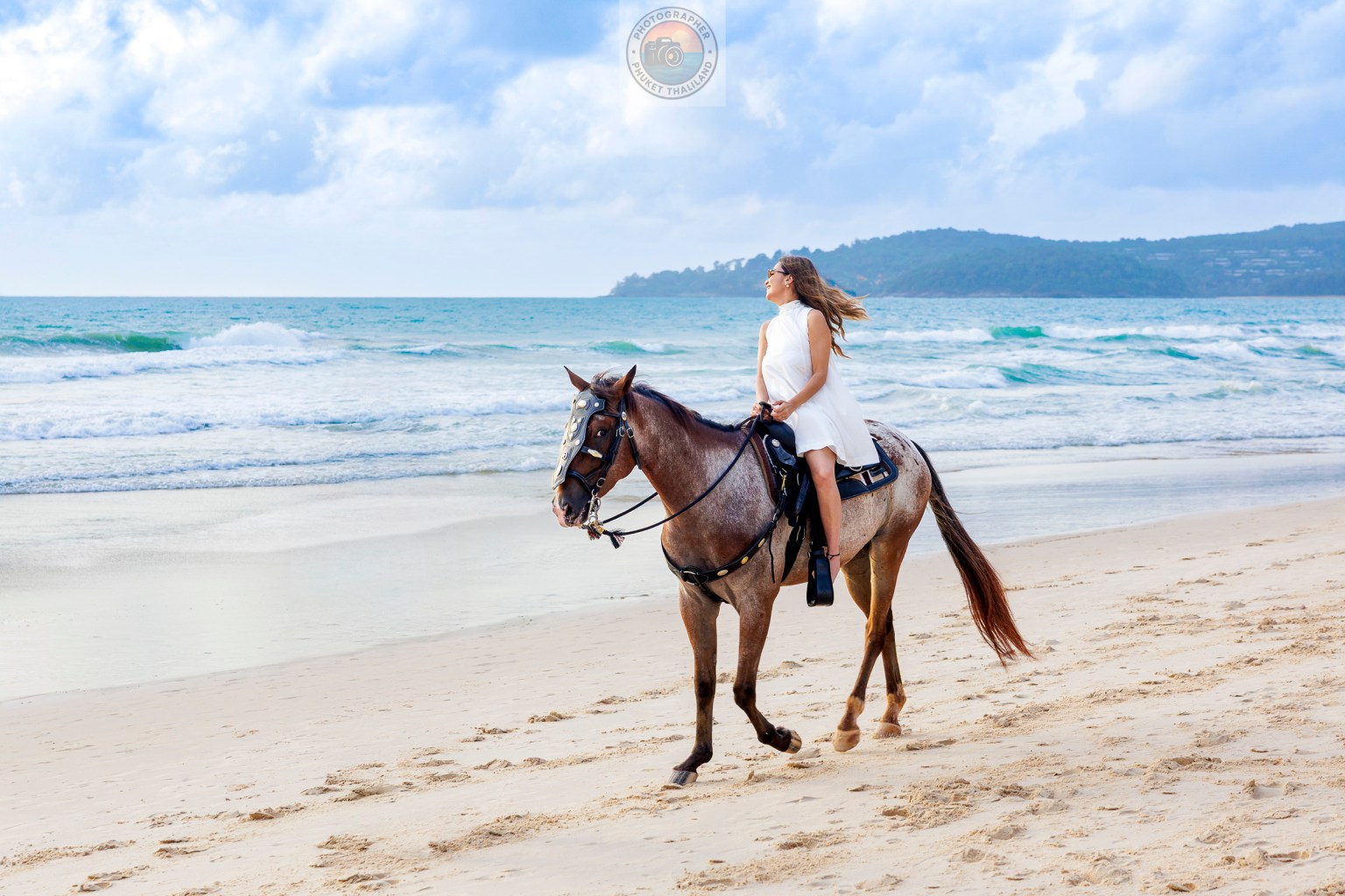 A woman in a white dress riding a horse along a sandy beach with waves in the background.