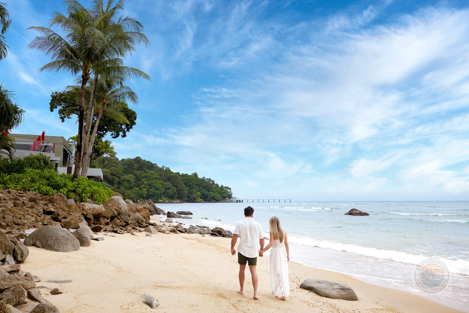 A couple walking hand in hand along a sandy beach, with palm trees and a scenic view of the sea and mountains in the background.