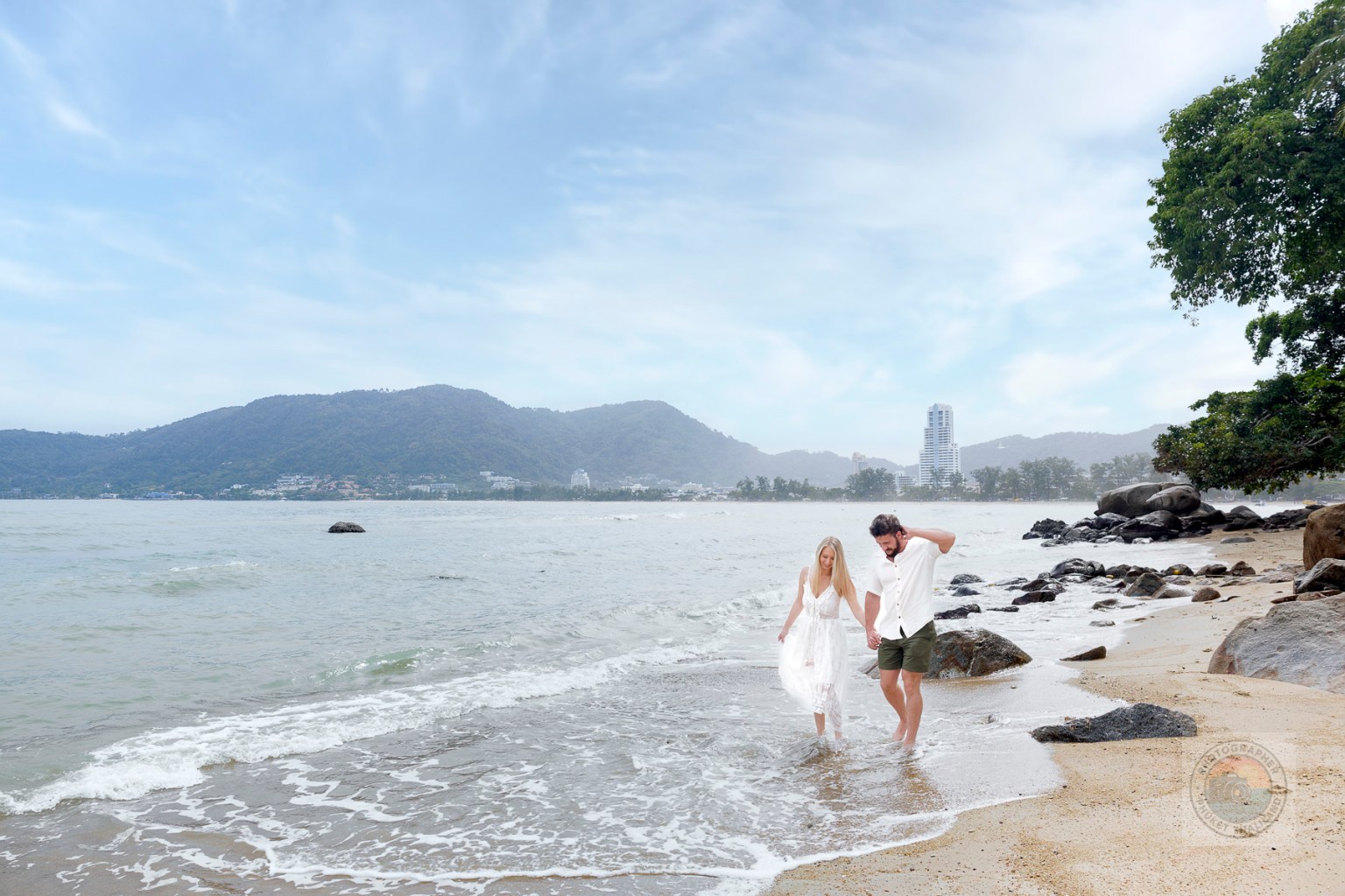 A couple walking hand in hand along a sandy beach, with gentle ocean waves lapping at their feet and mountains in the background.