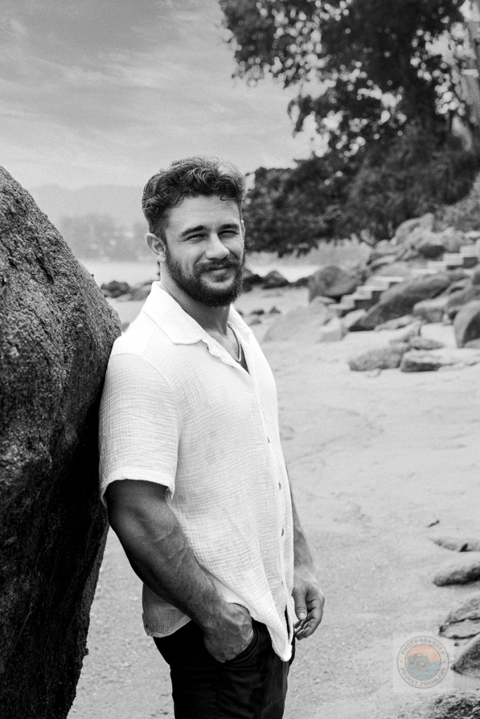 A man with curly hair and a beard stands confidently against a large rock on a sandy beach, wearing a white short-sleeve shirt and dark pants.