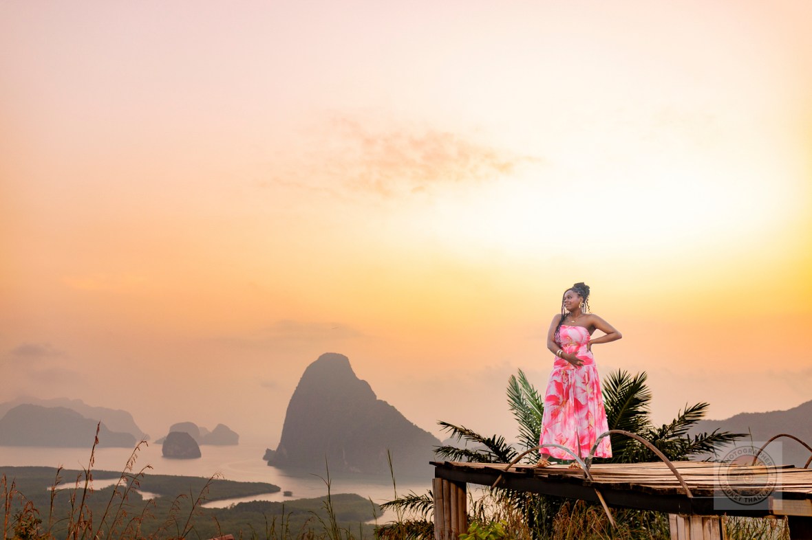 A woman in a pink floral dress stands on a wooden platform at Samet Nangshe viewpoint, with a breathtaking sunrise and limestone formations of Phang Nga Bay in the background.