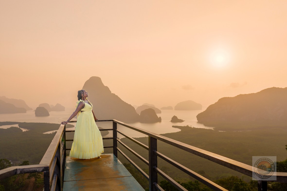 A woman in a yellow dress stands on a viewing platform at Samet Nangshe, overlooking the scenic limestone formations of Phang Nga Bay during sunrise.
