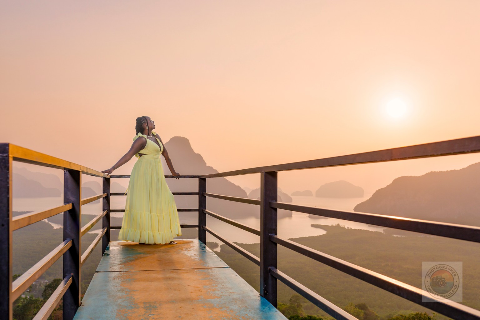 A woman in a yellow dress stands on a viewpoint railing at Samet Nangshe, overlooking the stunning limestone formations of Phang Nga Bay during sunrise.