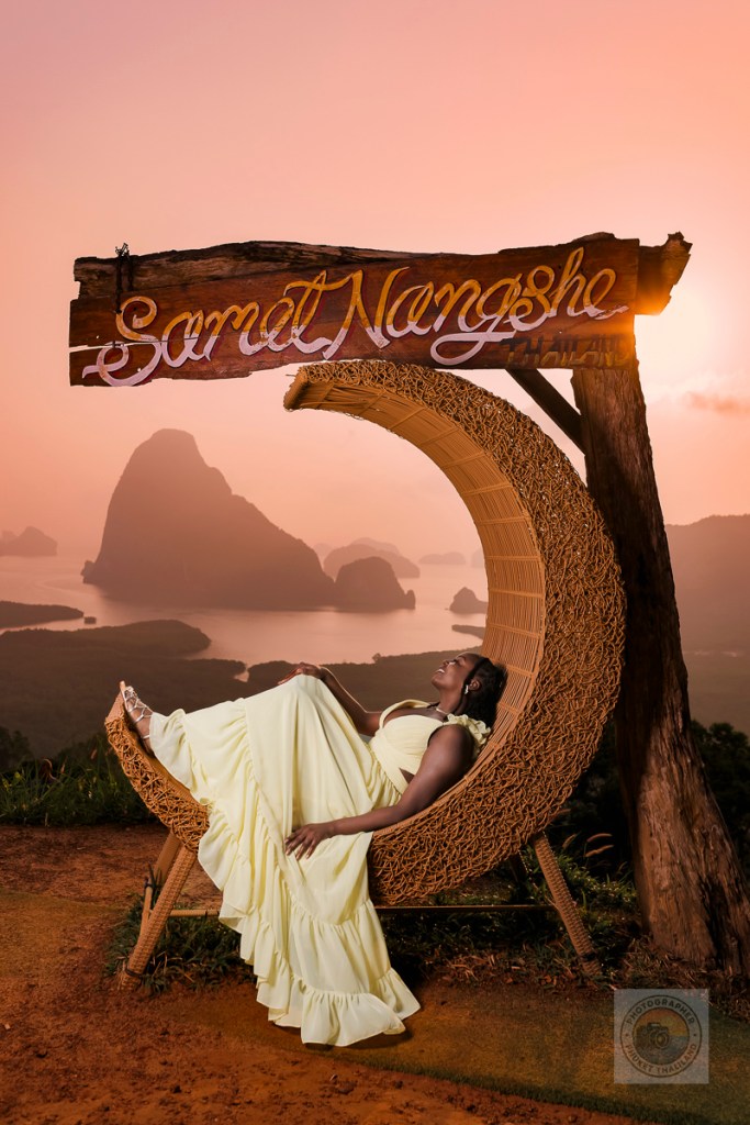 A woman in a flowing yellow dress reclining on a woven chair with the sign 'Samet Nangshe' in the background, overlooking a scenic view of limestone formations and a hazy sunrise.