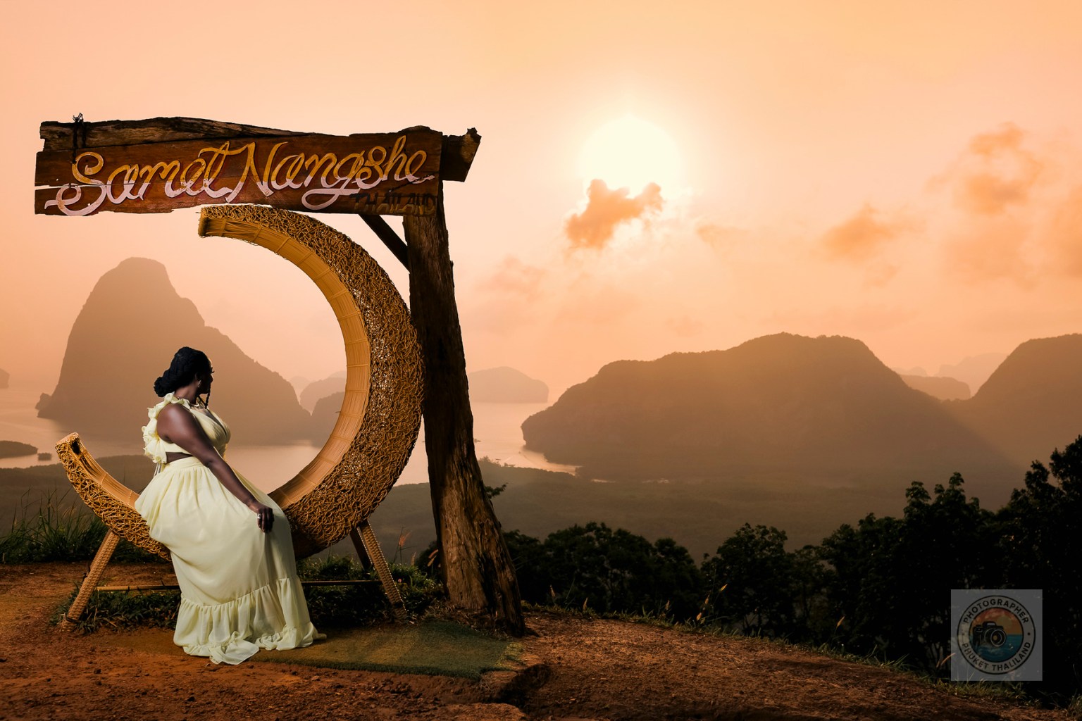 A woman in a light yellow dress sits on a decorative chair at the Samet Nangshe viewpoint, overlooking the scenic limestone formations of Phang Nga Bay at sunset.