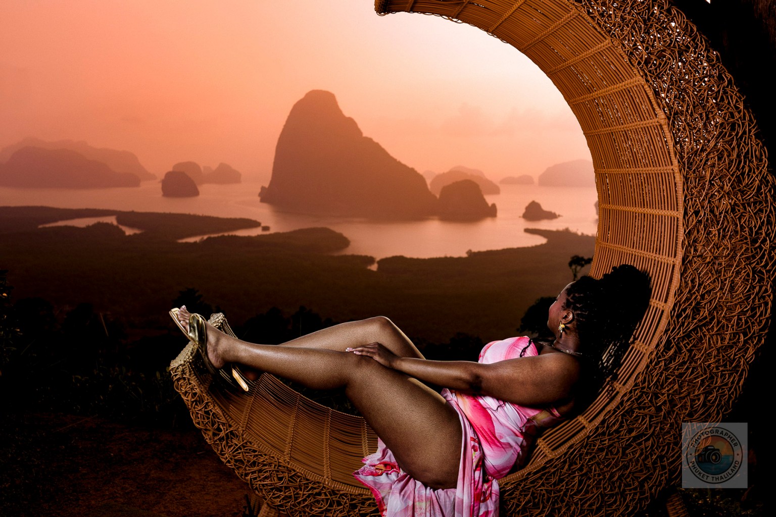 A woman relaxing in a round wicker chair, overlooking a stunning landscape of mountains and water during sunset.