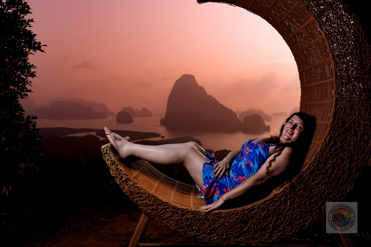 A woman in a blue floral dress reclining on a woven chair at sunset, with scenic mountains and water in the background.
