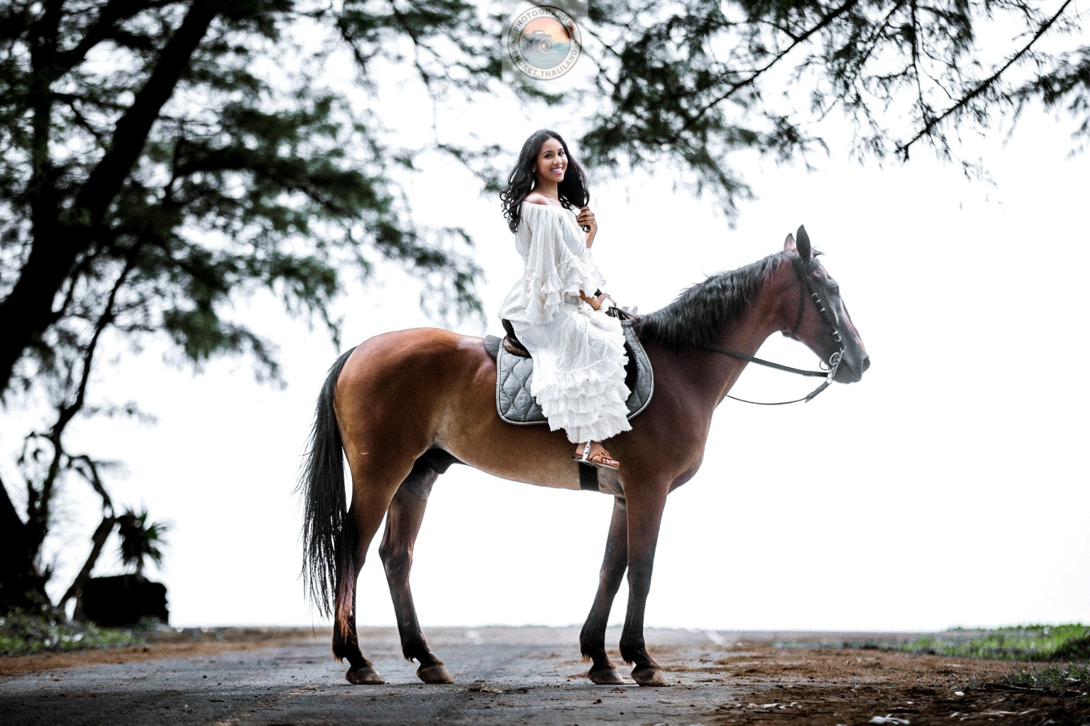 A young woman dressed in a flowing white dress poses gracefully on a brown horse, surrounded by lush greenery and an open road.
