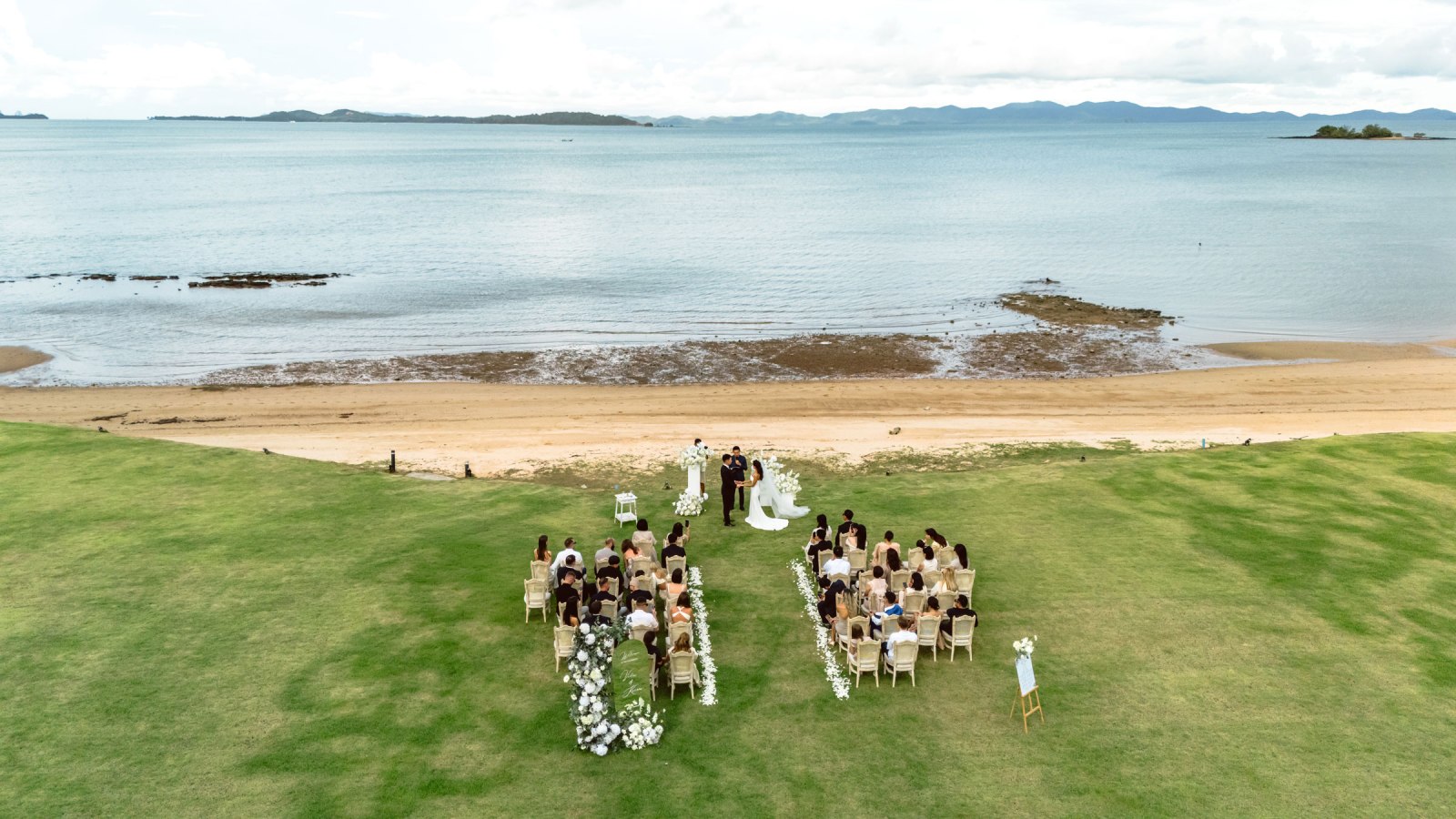 Aerial view of a wedding ceremony set on a beach with guests seated in rows on green grass, overlooking the ocean.